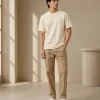 Man wearing a relaxed-fit cream t-shirt paired with khaki chinos and white sneakers, standing in a sunlit minimal interior.