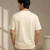 Man wearing a relaxed-fit cream t-shirt paired with khaki chinos and white sneakers, standing in a sunlit minimal interior.
