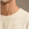 Man wearing a relaxed-fit cream t-shirt paired with khaki chinos and white sneakers, standing in a sunlit minimal interior.