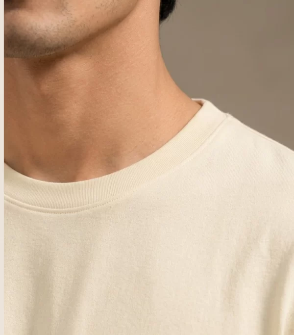 Man wearing a relaxed-fit cream t-shirt paired with khaki chinos and white sneakers, standing in a sunlit minimal interior.