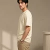 Man wearing a relaxed-fit cream t-shirt paired with khaki chinos and white sneakers, standing in a sunlit minimal interior.