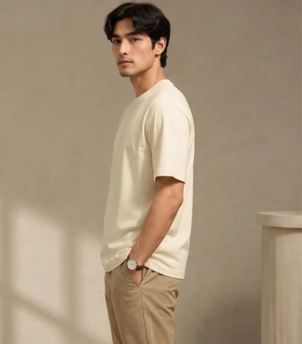 Man wearing a relaxed-fit cream t-shirt paired with khaki chinos and white sneakers, standing in a sunlit minimal interior.