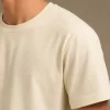 Man wearing a relaxed-fit cream t-shirt paired with khaki chinos and white sneakers, standing in a sunlit minimal interior.