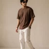 Man wearing brown oversized t-shirt with cream cargo pants, sunglasses, and white sneakers in a minimal studio setting.