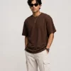 Man wearing brown oversized t-shirt with cream cargo pants, sunglasses, and white sneakers in a minimal studio setting.