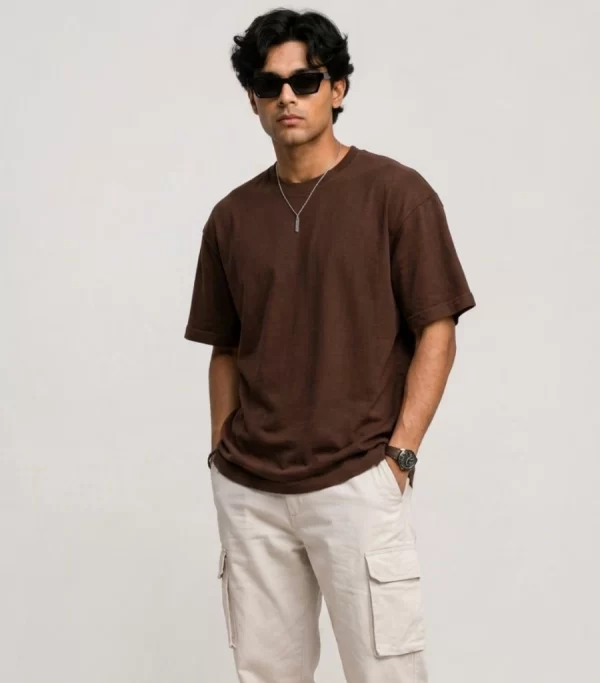 Man wearing brown oversized t-shirt with cream cargo pants, sunglasses, and white sneakers in a minimal studio setting.
