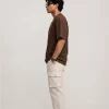 Man wearing brown oversized t-shirt with cream cargo pants, sunglasses, and white sneakers in a minimal studio setting.