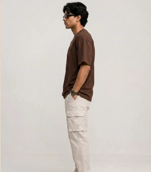 Man wearing brown oversized t-shirt with cream cargo pants, sunglasses, and white sneakers in a minimal studio setting.