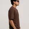 Man wearing brown oversized t-shirt with cream cargo pants, sunglasses, and white sneakers in a minimal studio setting.