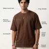 Man wearing brown oversized t-shirt with cream cargo pants, sunglasses, and white sneakers in a minimal studio setting.